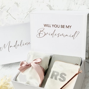 May include: White gift box with a pink ribbon and a white bag with a pink ribbon. The box has a white lid with the text "Will you be my Bridesmaid?" in brown lettering. The box also has a white label with the name "Madeline" in black lettering. The box contains a white pouch with the word "BRIDE" in silver glitter letters.