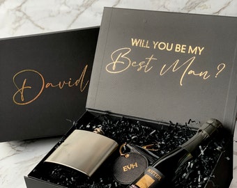 May include: A black gift box with gold lettering that says "Will you be my Best Man?" The box is open and contains a silver flask, a leather keychain with the initials "EVH", and a bottle of prosecco.