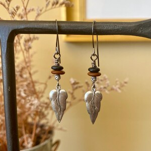 May include: Silver leaf dangle earrings with brown and black beads.  The earrings feature textured silver leaf charms suspended from delicate silver hooks. Three small beads in brown and black are positioned above each leaf. These stylish earrings are perfect for adding a touch of nature-inspired elegance to any outfit.