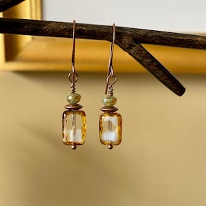 Peut inclure: Une paire de boucles d'oreilles pendantes avec un fil de cuivre et une perle de verre ambrée rectangulaire avec des tourbillons blancs. La perle est surmontée d'une plus petite perle de verre verte.