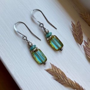 May include: A pair of handcrafted earrings with silver-tone ear wires. Each earring features a rectangular, teal-colored glass bead with yellow edges, accented by turquoise and silver beads. The earrings are displayed on a white surface with dried plants.