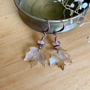 May include: A pair of copper wire earrings with a delicate, translucent, light brown leaf charm and a light pink glass bead on each earring.