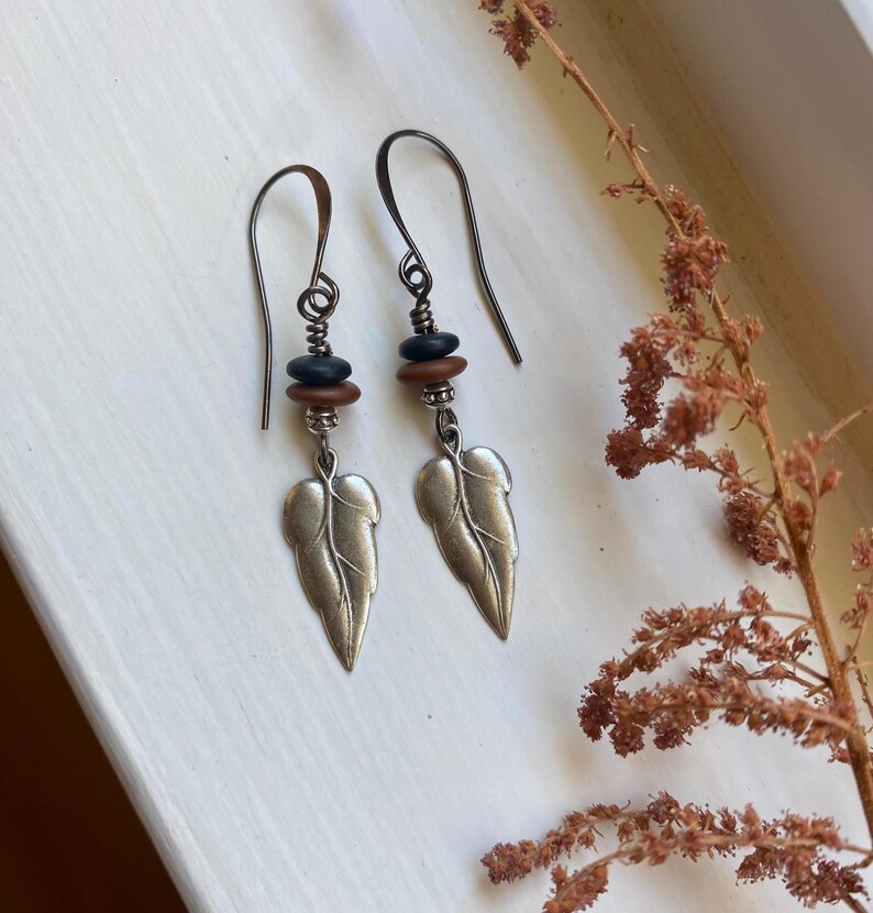 May include: A pair of silver leaf earrings with brown and black beads. The earrings are hanging from silver hooks.