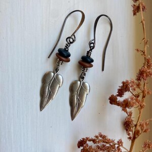 May include: Silver leaf earrings with brown and black beads hanging from a silver hook.