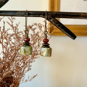 Puede incluir: Un par de pendientes colgantes con una cuenta rectangular de tonos verdes y beige jaspeados, suspendida de un gancho de metal oscuro. Sobre la cuenta principal, un disco met&aacute;lico y una cuenta roja facetada con alambre.