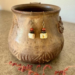 Puede incluir: Un par de pendientes colgantes con ganchos de bronce, peque&ntilde;as cuentas rojas facetadas, espaciadores de metal oscuro y cuentas rectangulares de cer&aacute;mica color hueso con un sutil veteado marr&oacute;n sobre una taza.