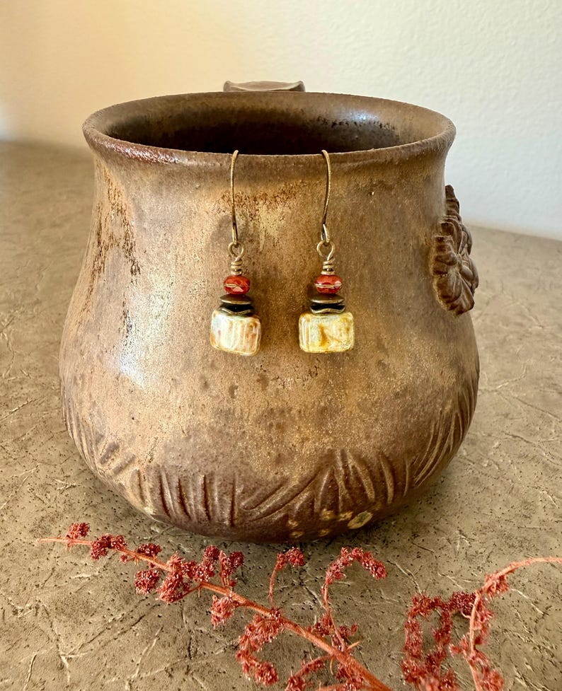 Puede incluir: Un par de pendientes colgantes con ganchos de bronce, peque&ntilde;as cuentas rojas facetadas, espaciadores de metal oscuro y cuentas rectangulares de cer&aacute;mica color hueso con un sutil veteado marr&oacute;n sobre una taza.