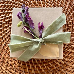 Puede incluir: Una caja de regalo cuadrada de cart&oacute;n marr&oacute;n claro atada con una cinta deshilachada de color verde salvia. Una ramita de flores secas moradas y un peque&ntilde;o capullo verde p&aacute;lido est&aacute;n sujetos bajo el nudo, junto a una etiqueta de papel en forma de coraz&oacute;n.