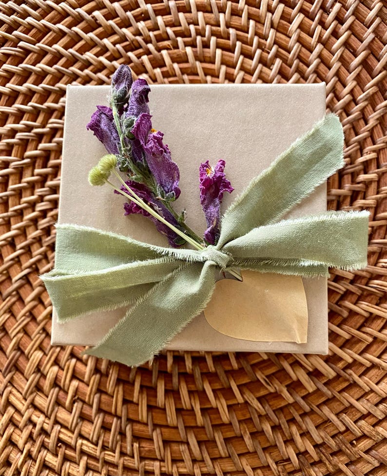 Puede incluir: Una caja de regalo cuadrada de cart&oacute;n marr&oacute;n claro atada con una cinta deshilachada de color verde salvia. Una ramita de flores secas moradas y un peque&ntilde;o capullo verde p&aacute;lido est&aacute;n sujetos bajo el nudo, junto a una etiqueta de papel en forma de coraz&oacute;n.