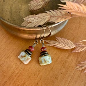 Puede incluir: Un par de pendientes colgantes con ganchos de bronce, peque&ntilde;as cuentas esf&eacute;ricas rojas y colgantes rectangulares de estilo cer&aacute;mico con un esmalte moteado en tonos crema, beige y marr&oacute;n. Cuelgan del borde de un cuenco met&aacute;lico.