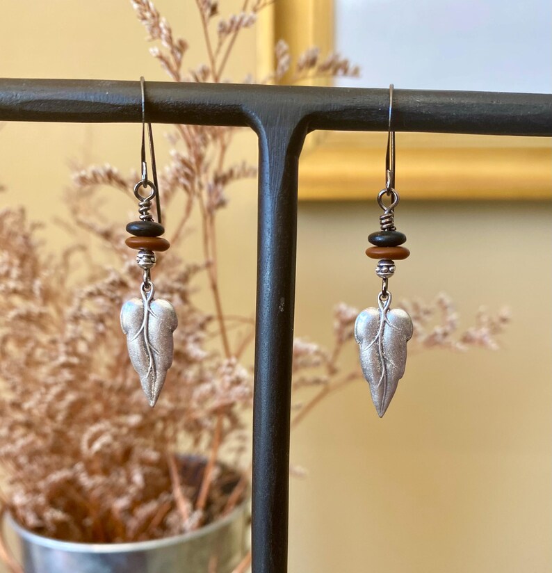 May include: Silver leaf earrings with brown and black beads hanging from a black metal stand.