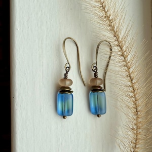 May include: A pair of handmade earrings with a vintage aesthetic. Each earring features a blue, rectangular bead with a frosted finish, complemented by a light brown bead and antique brass-colored metal accents. The earrings hang from curved ear wires.