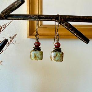 Puede incluir: Un par de pendientes colgantes con una cuenta rectangular de color verde y marr&oacute;n jaspeado en la base, rematada con un peque&ntilde;o disco de metal color bronce y una cuenta roja transl&uacute;cida facetada, suspendidos de un gancho de metal color bronce.