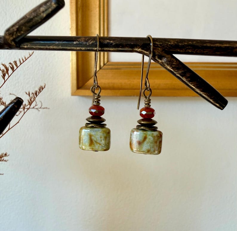 Puede incluir: Un par de pendientes colgantes con una cuenta rectangular de color verde y marr&oacute;n jaspeado en la base, rematada con un peque&ntilde;o disco de metal color bronce y una cuenta roja transl&uacute;cida facetada, suspendidos de un gancho de metal color bronce.