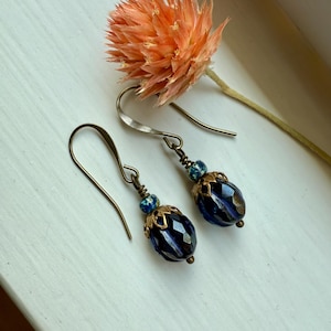 May include: A pair of handcrafted earrings with dark blue faceted beads and gold-coloured filigree caps. The earrings have a vintage aesthetic, with a hook closure. An orange dried flower is in the background.