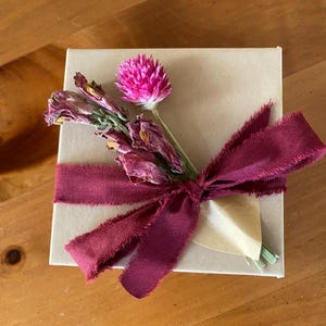 May include: A small gift box wrapped in brown paper with a burgundy ribbon and a bouquet of dried flowers, including a pink flower.