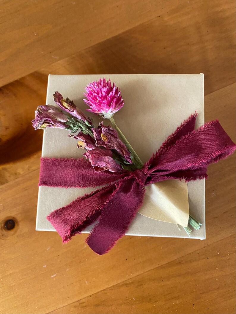 May include: A small gift box wrapped in brown paper with a burgundy ribbon and a bouquet of dried flowers, including a pink flower.