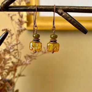 May include: A pair of dangle earrings with a square amber-colored glass bead and a smaller green bead above it. The earrings are made with copper wire.