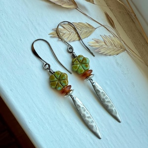 May include: A pair of handcrafted earrings featuring a green flower-shaped bead, an orange bead, and a silver, textured, elongated teardrop pendant. The earrings have a silver hook and are displayed on a white surface with dried wheat stalks.