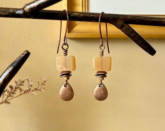 Puede incluir: Pendientes hechos a mano con una cuenta cuadrada de color melocotón claro, un disco de color cobre y un dije en forma de lágrima. Los pendientes cuelgan de alambres para las orejas de color cobre. Las joyas se exhiben en una rama marrón oscuro.