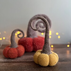 May include: Three crocheted pumpkins in different colors: red, orange, and yellow. The pumpkins have a curly stem and are sitting on a wooden surface.