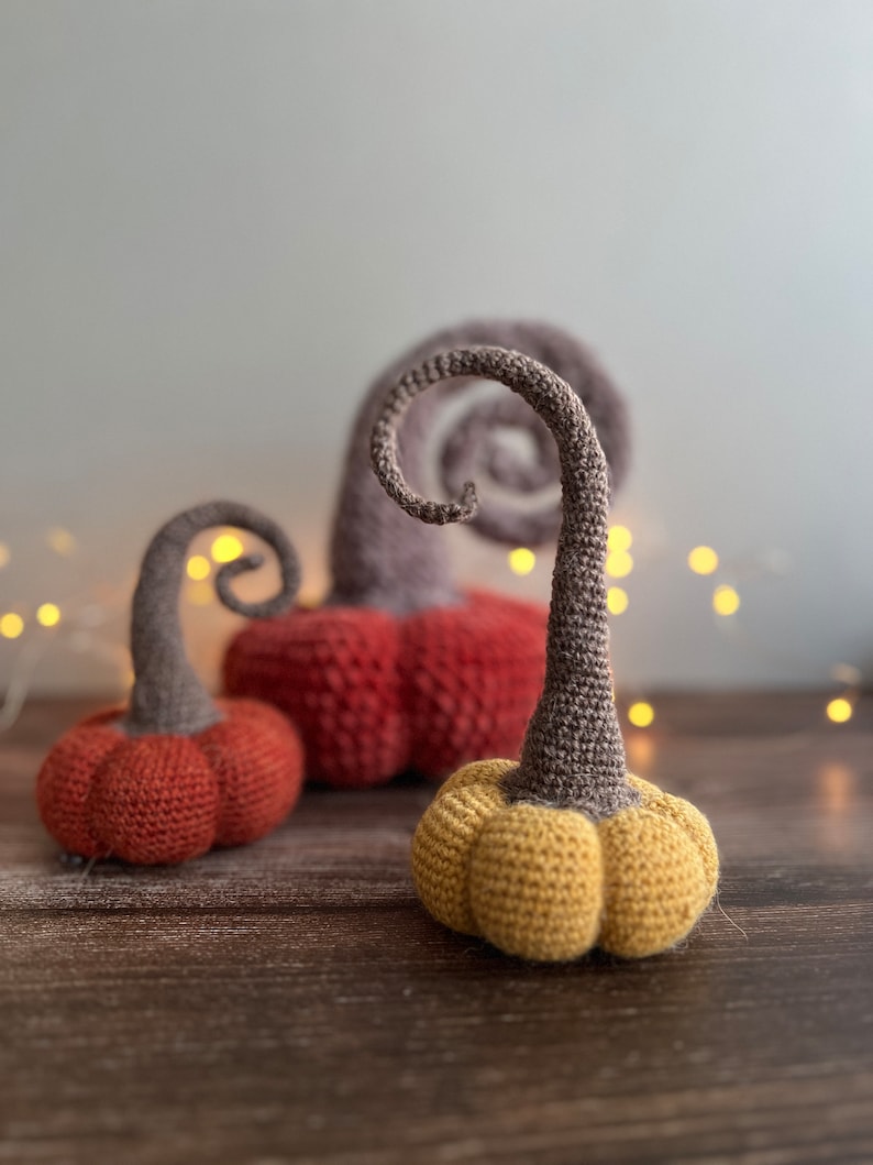 May include: Three crocheted pumpkins in different colors: red, orange, and yellow. The pumpkins have a curly stem and are sitting on a wooden surface.