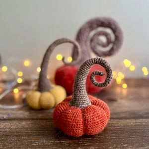 May include: Three crocheted pumpkins in orange, yellow, and brown. The pumpkins have a spiral stem and are sitting on a wooden surface.