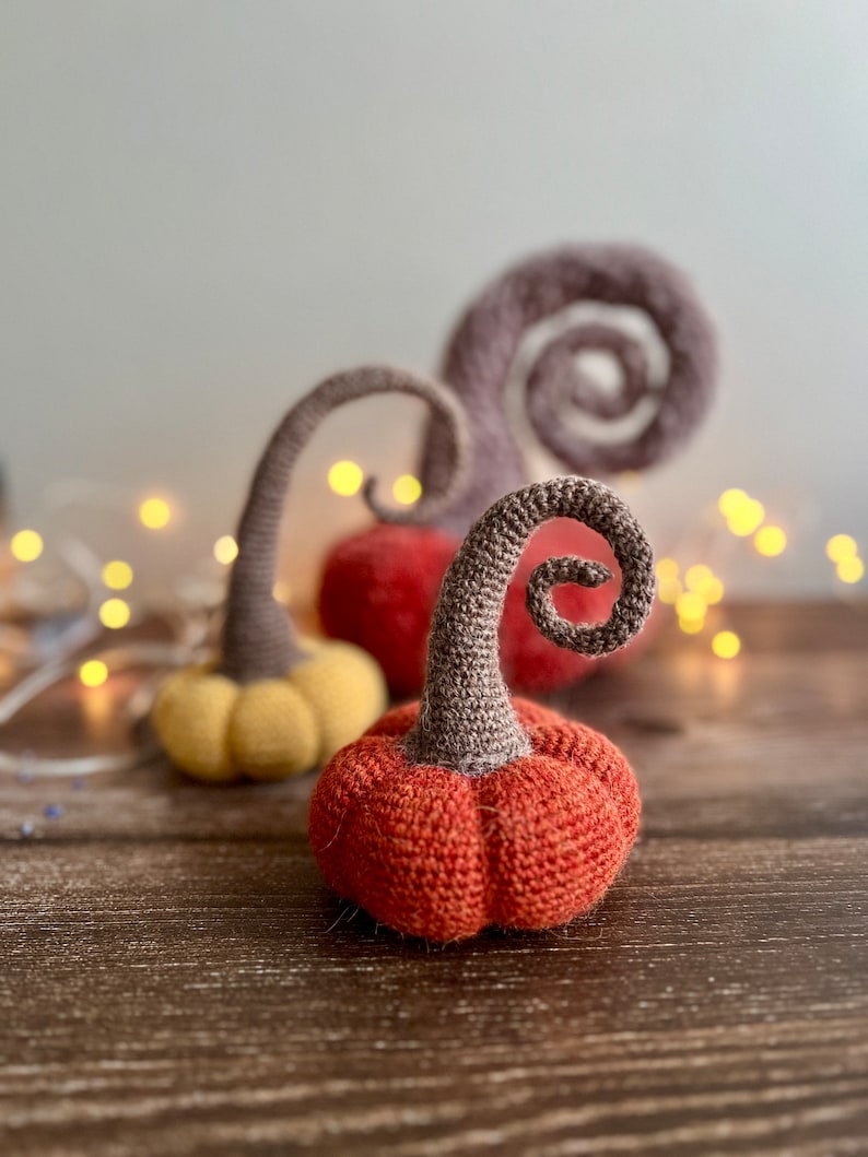 May include: Three crocheted pumpkins in orange, yellow, and brown. The pumpkins have a spiral stem and are sitting on a wooden surface.