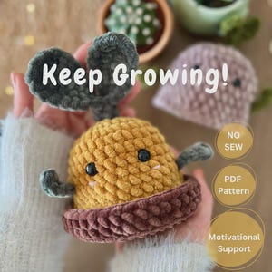 Puede incluir: Una planta de ganchillo amarilla y marrón hecha a mano con el texto "Keep Growing!". La planta amigurumi tiene una cara sonriente, hojas verdes y una maceta marrón. La imagen también incluye una pequeña planta de ganchillo rosa y una suculenta.