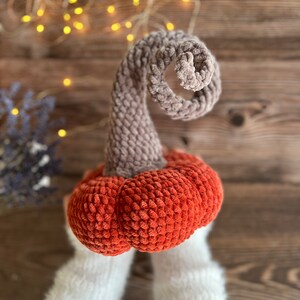 May include: A crocheted pumpkin with a brown stem and an orange body. The pumpkin is held by two hands wearing white knitted gloves.