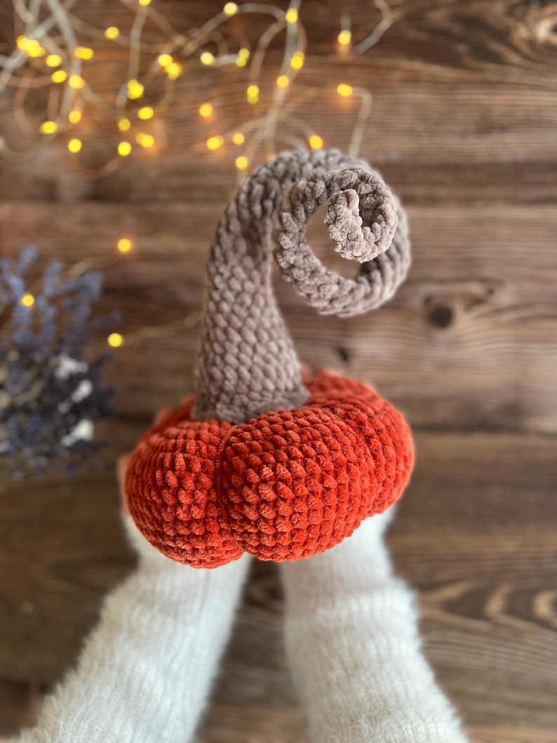 May include: A crocheted pumpkin with a brown stem and an orange body. The pumpkin is held by two hands wearing white knitted gloves.