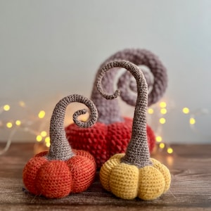 May include: Three crocheted pumpkins in different sizes and colors. The largest pumpkin is red, the middle pumpkin is orange, and the smallest pumpkin is yellow. All three pumpkins have brown stems.