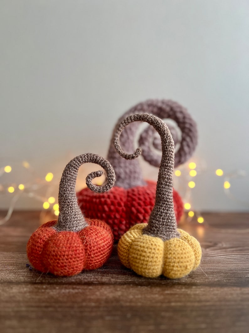 May include: Three crocheted pumpkins in different sizes and colors. The largest pumpkin is red, the middle pumpkin is orange, and the smallest pumpkin is yellow. All three pumpkins have brown stems.