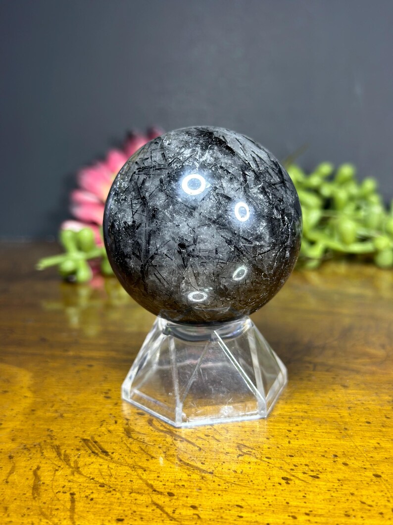 Może przedstawiać: Czarna kula z turmalinu z misterynymi wzorami wewnętrznymi, spoczywająca na przezroczystym, fasetowanym kryształowym stojaku. Kula ma wypolerowaną powierzchnię z czarnymi i szarymi smugami. Stojak jest sześciokątny i przezroczysty, eksponując szczeg&oacute;ły.