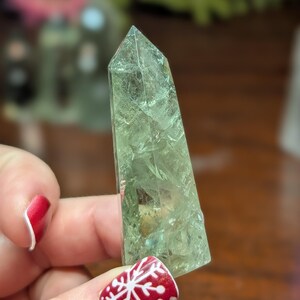 May include: A light green crystal obelisk with a pointed top. The crystal has internal patterns and inclusions. It is held by a hand with red nail polish and a snowflake design on the nail.