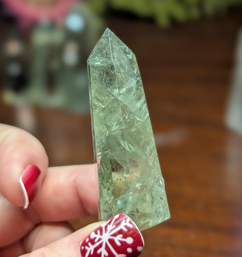 May include: A light green crystal obelisk with a pointed top. The crystal has internal patterns and inclusions. It is held by a hand with red nail polish and a snowflake design on the nail.