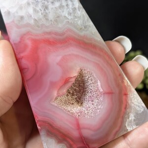 May include: A polished, rectangular agate stone with a striking pattern of pink, red, and white bands. The stone features a central geode cavity with a crystalline interior. The overall aesthetic is natural and decorative.