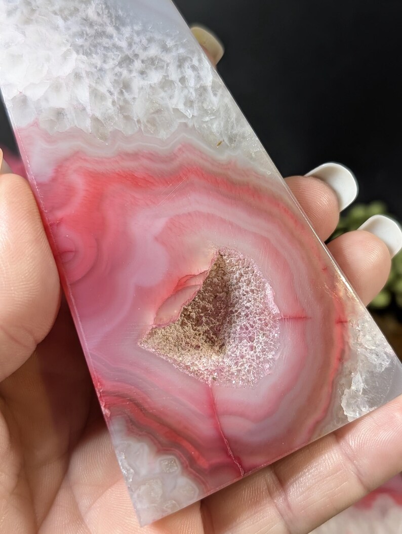 May include: A polished, rectangular agate stone with a striking pattern of pink, red, and white bands. The stone features a central geode cavity with a crystalline interior. The overall aesthetic is natural and decorative.