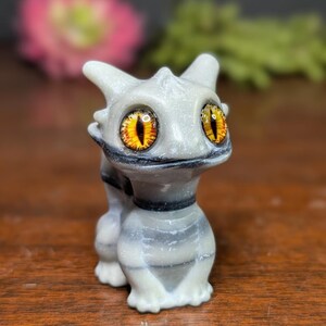 Pode incluir: Uma pequena figura cinza claro de uma criatura semelhante a um drag&atilde;o com grandes olhos amarelos e laranja. A figura tem um corpo listrado e uma coleira preta. O fundo est&aacute; desfocado, com toques de rosa e verde.