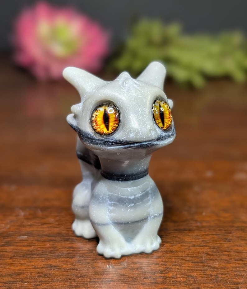 Pode incluir: Uma pequena figura cinza claro de uma criatura semelhante a um drag&atilde;o com grandes olhos amarelos e laranja. A figura tem um corpo listrado e uma coleira preta. O fundo est&aacute; desfocado, com toques de rosa e verde.