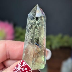 May include: A clear, faceted crystal point with a pale green and iridescent sheen. The crystal is held in a hand with a red and white snowflake design on the fingernail. The crystal is a natural, polished stone.