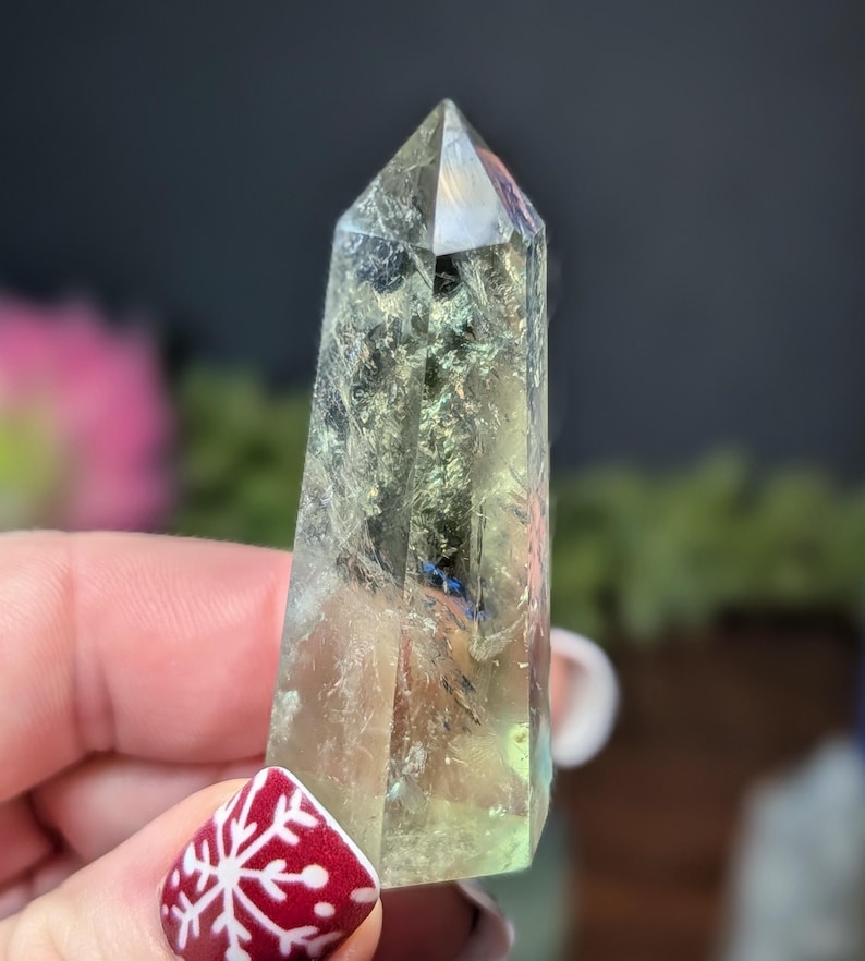 May include: A clear, faceted crystal point with a pale green and iridescent sheen. The crystal is held in a hand with a red and white snowflake design on the fingernail. The crystal is a natural, polished stone.