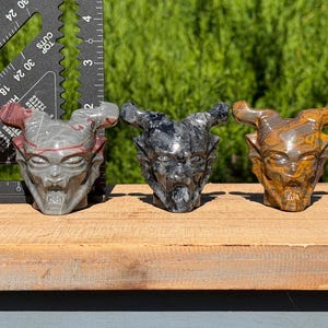 May include: Five carved stone demon head figurines arranged in a row, featuring varied colors including grey, black, brown, and white. A black triangular ruler is positioned behind the first figurine, showing measurements in inches.