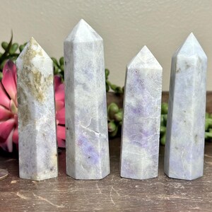 May include: Four white and purple crystal points, also known as wands or towers, are arranged in a row on a wooden surface. The crystals are all different sizes and have a natural, rough texture.