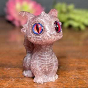 Pode incluir: Uma pequena estatueta de drag&atilde;o cor de lavanda com olhos grandes e detalhados. Os olhos s&atilde;o uma combina&ccedil;&atilde;o marcante de vermelho, preto e azul. O drag&atilde;o est&aacute; em p&eacute;, com a cauda enrolada atr&aacute;s de si. A estatueta &eacute; feita de um material salpicado.