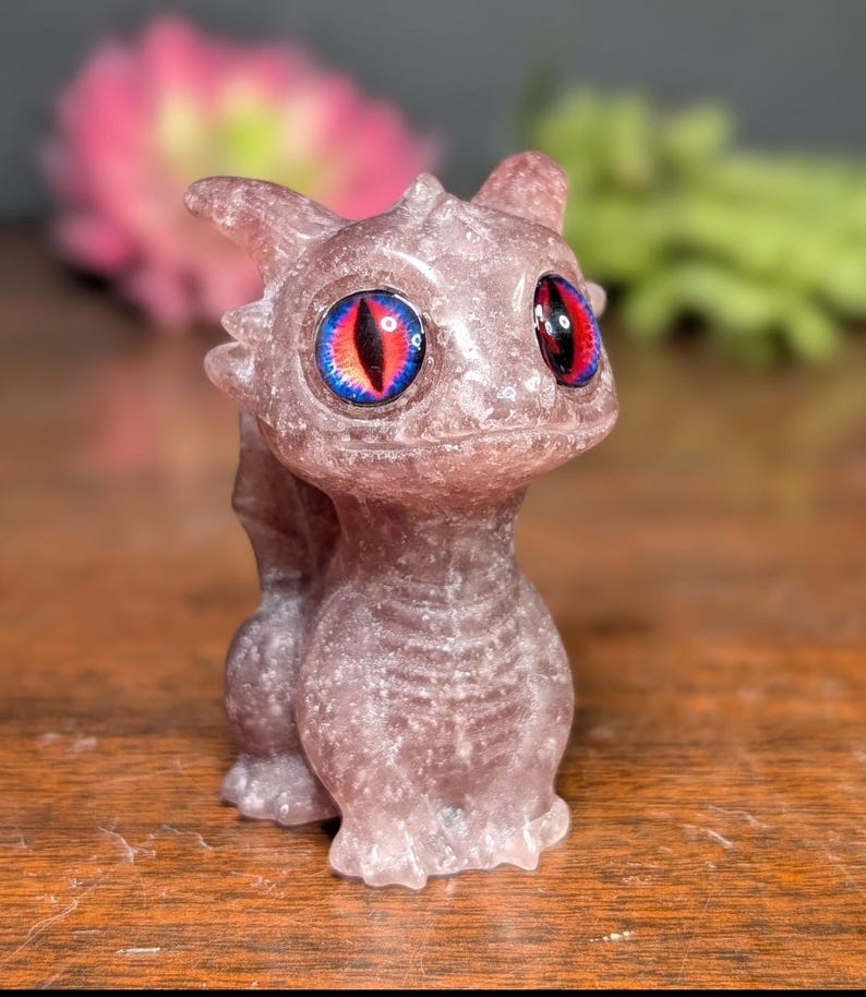Pode incluir: Uma pequena estatueta de drag&atilde;o cor de lavanda com olhos grandes e detalhados. Os olhos s&atilde;o uma combina&ccedil;&atilde;o marcante de vermelho, preto e azul. O drag&atilde;o est&aacute; em p&eacute;, com a cauda enrolada atr&aacute;s de si. A estatueta &eacute; feita de um material salpicado.