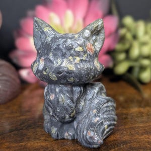 May include: A carved stone cat figurine in shades of gray, black, and green, with flecks of orange and yellow. The cat is sitting upright with its ears perked up. The background is blurred, with pink and green flowers.