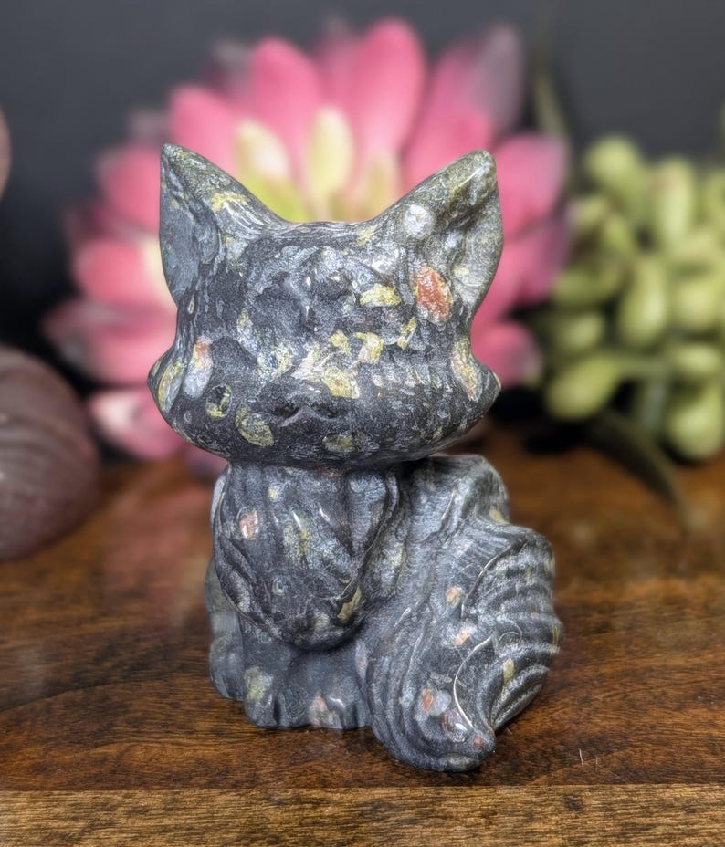 May include: A carved stone cat figurine in shades of gray, black, and green, with flecks of orange and yellow. The cat is sitting upright with its ears perked up. The background is blurred, with pink and green flowers.