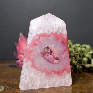 May include: A polished, pink and white agate crystal obelisk. The crystal features a central geode with a deep pink hue, surrounded by concentric bands of pink and white. The obelisk is a natural stone with a smooth, polished surface.