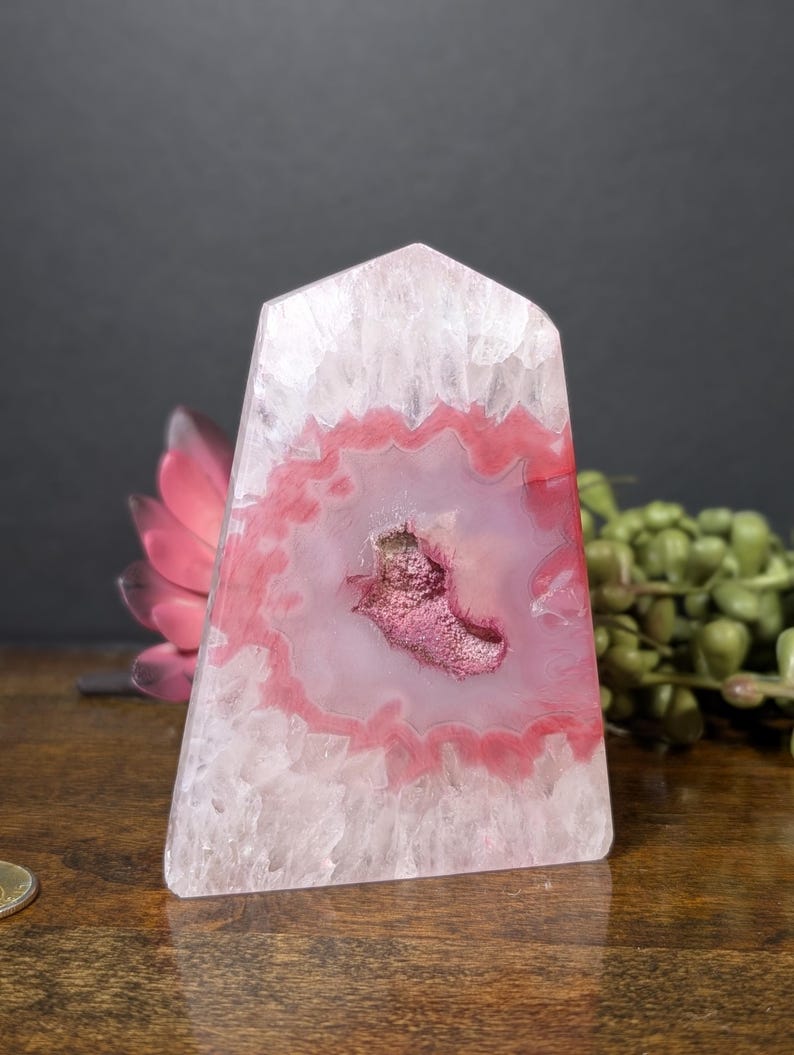 May include: A polished, pink and white agate crystal obelisk. The crystal features a central geode with a deep pink hue, surrounded by concentric bands of pink and white. The obelisk is a natural stone with a smooth, polished surface.
